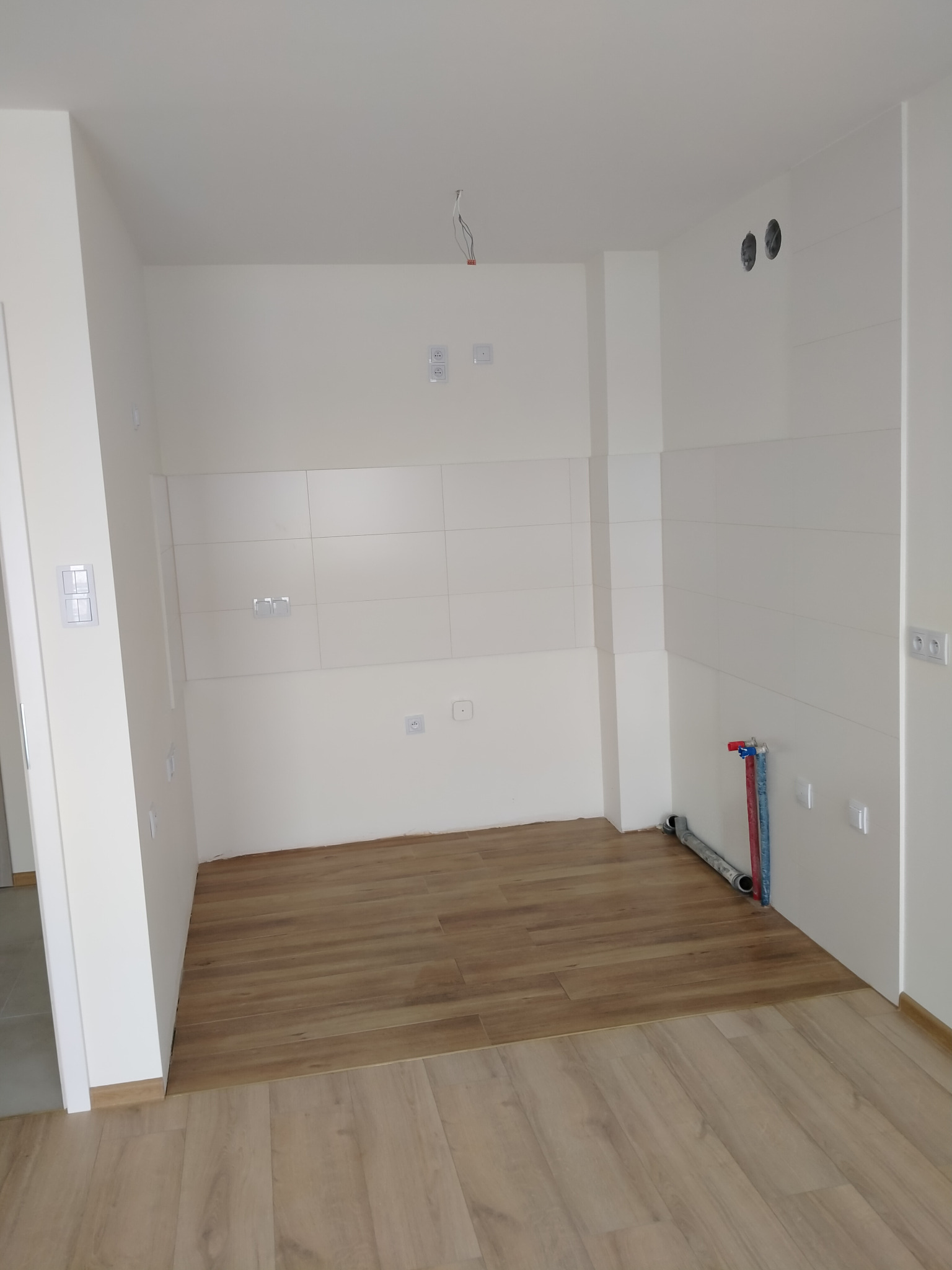 Wnętrze kuchni w stanie deweloperskim: białe ściany z gładkimi płytkami, drewnopodobne panele podłogowe, widoczne instalacje wodne i elektryczne, przygotowane do dalszych prac wykończeniowych.