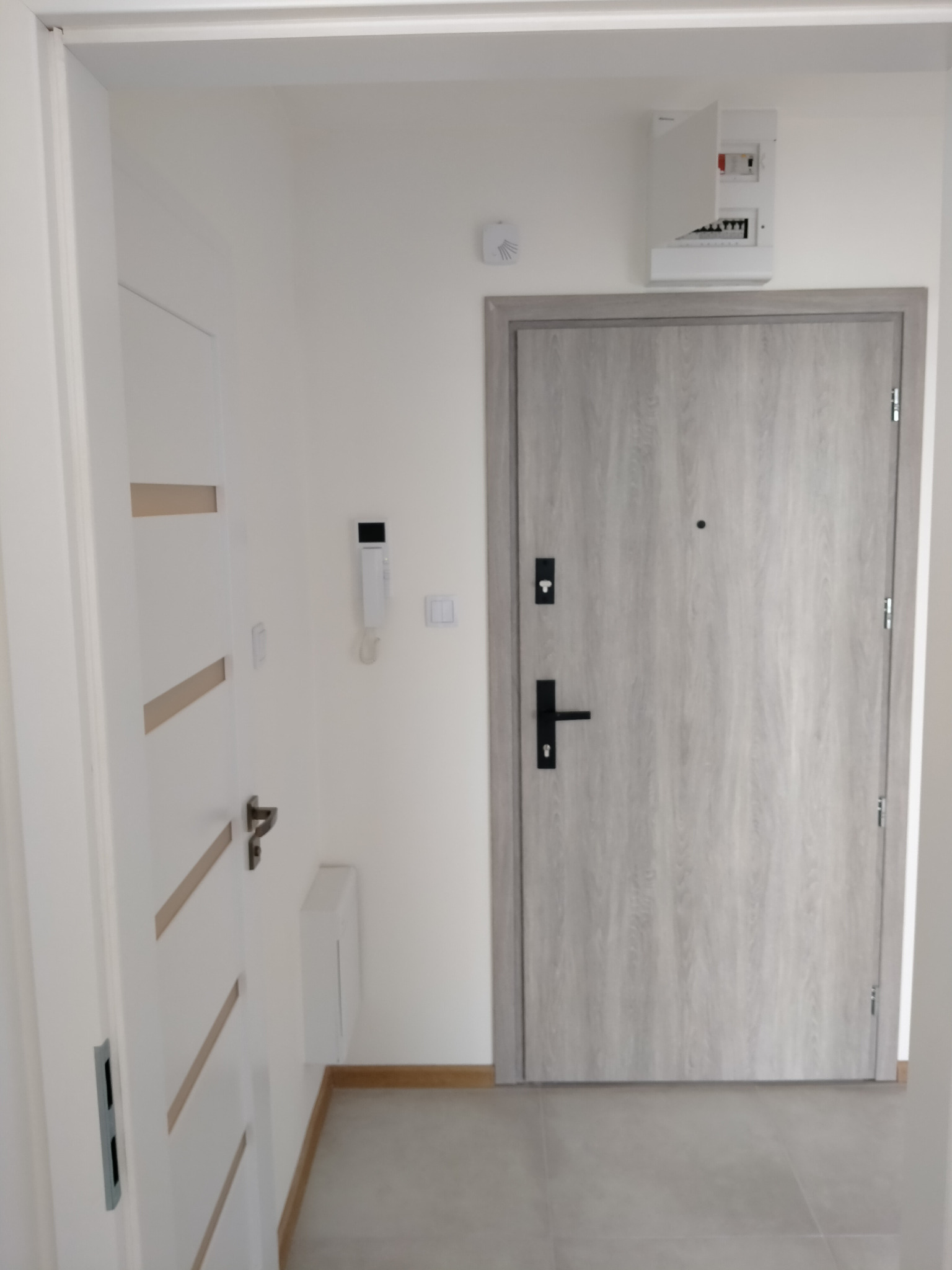 Nowoczesny przedpokój z szarą, drewnopodobną okleiną na drzwiach wejściowych, białe drzwi wewnętrzne z poziomymi wstawkami i szarymi panelami podłogowymi.