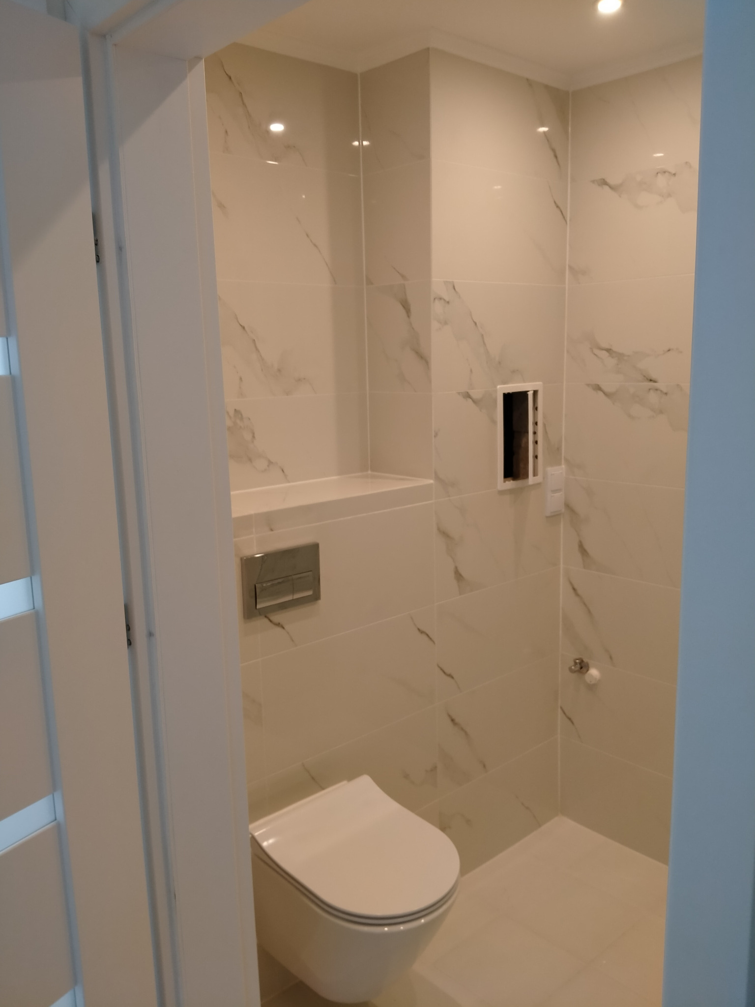 Nowoczesna łazienka po remoncie: białe płytki imitujące marmur, podtynkowa toaleta, zabudowana półka i wnęka na kosmetyki, chromowany przycisk spłuczki.