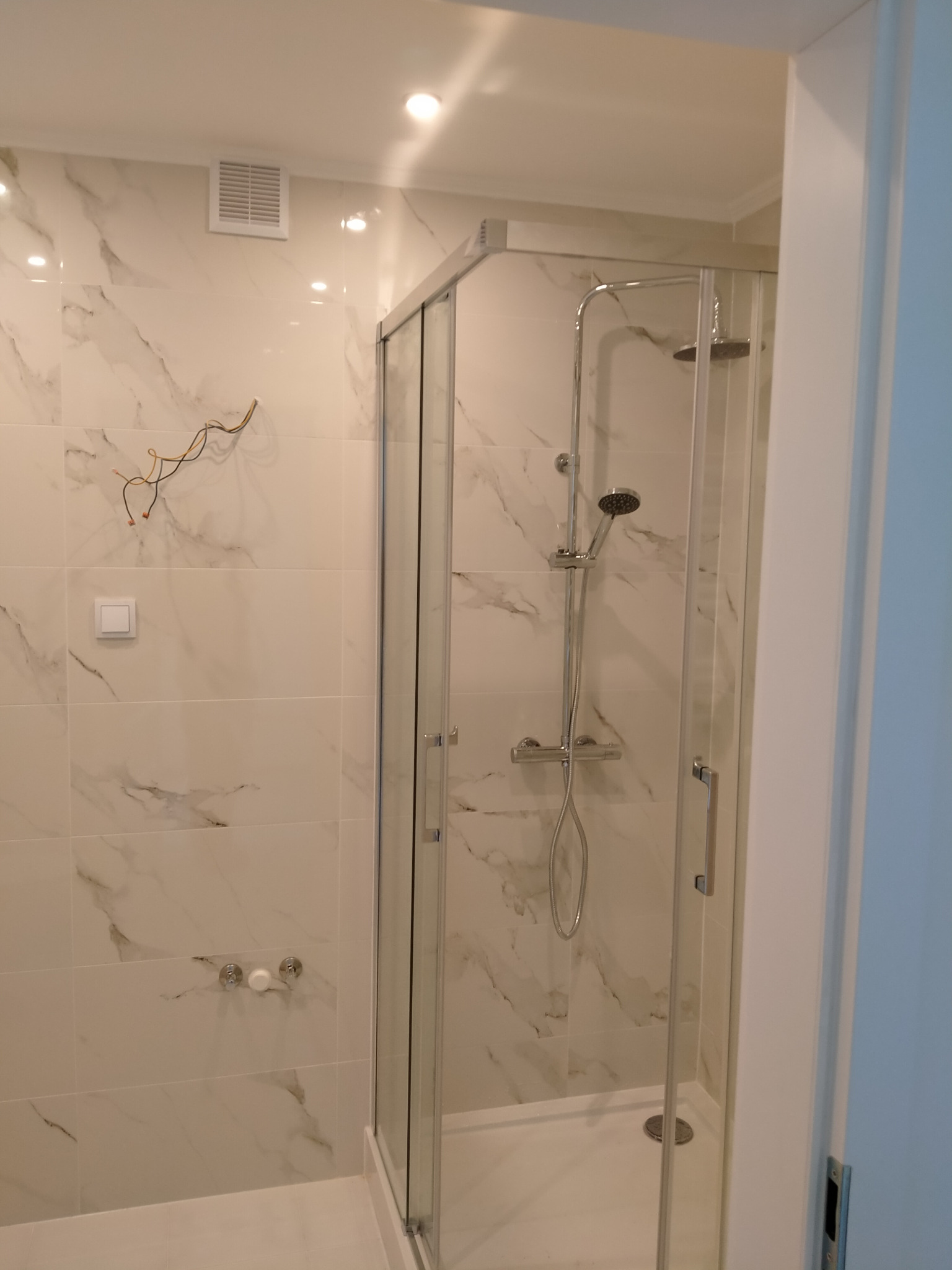 Nowoczesna kabina prysznicowa z chromowaną armaturą na tle białych płytek imitujących marmur, widoczne wystające przewody elektryczne i włącznik światła.