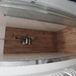 Wykończona kabina prysznicowa z brązowymi płytkami imitującymi drewno i odpływem liniowym. Widoczne białe płytki na dole i sufit z białych kwadratowych płytek.