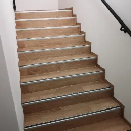 Schody wewnętrzne wykończone panelami drewnopodobnymi z aluminiowymi listwami antypoślizgowymi i czarną metalową poręczą przy białej ścianie.