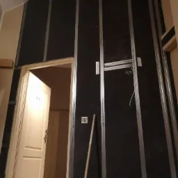 Montaż płyty karton gips na ścianie szkieletowej z metalowymi profilami, widoczne instalacje elektryczne i otwarta biała stolarka drzwiowa.