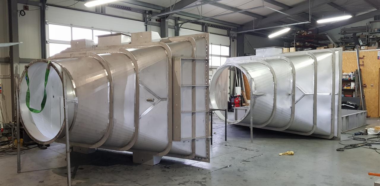 Dwie duże, metalowe konstrukcje o przekroju okrągłym i kwadratowym w hali w Sielecu, prawdopodobnie elementy instalacji wentylacyjnej lub przemysłowej, w trakcie montażu lub przygotowania...