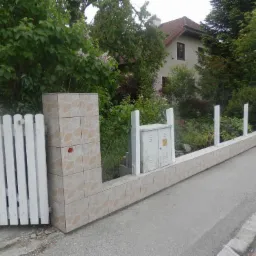 Oklejone płytkami ogrodzenie z białymi sztachetami i skrzynką elektryczną na tle zieleni i domu.