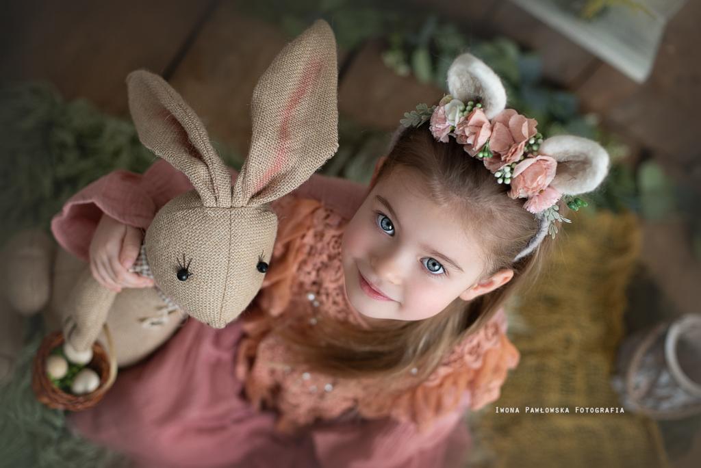Dziewczynka z niebieskimi oczami, w różowej sukience i opasce z kwiatami i uszami królika, trzymająca pluszowego królika z koszyczkiem jajek, widziana z góry.