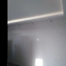 Biała ściana z cegły, oświetlenie punktowe sufitowe i podświetlany sufit, wykończona podłoga z paneli w odcieniach brązu, widoczne gniazdko elektryczne.