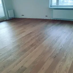 Wykończona podłoga z parkietu w jasnym odcieniu dębu, widoczne listwy przypodłogowe w trakcie montażu, ściany pomalowane na biało, z widocznymi gniazdkami elektrycznymi i grzejnikiem.