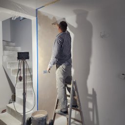 SNA BUD - Pracownik na drabinie aplikuje gładź na ścianę w jasnym wnętrzu. Widoczne schody w surowym stanie i narzędzia pracy. Ściana zabezpieczona taśmą malarską.