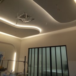 SNA BUD - Nowoczesny sufit podwieszany z oświetleniem LED w kształcie fal, widok z dołu. W tle ściana i okno. Na pierwszym planie rusztowanie i drabina.