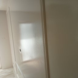 SNA BUD - Biała, świeżo pomalowana ściana z dekoracyjnymi listwami, widoczna pod lekkim kątem. Na podłodze folia ochronna. Gładka powierzchnia, odbijające światło wykończenie.