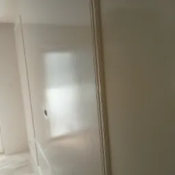 Biała, świeżo pomalowana ściana z dekoracyjnymi listwami, widoczna pod lekkim kątem. Na podłodze folia ochronna. Gładka powierzchnia, odbijające światło wykończenie.