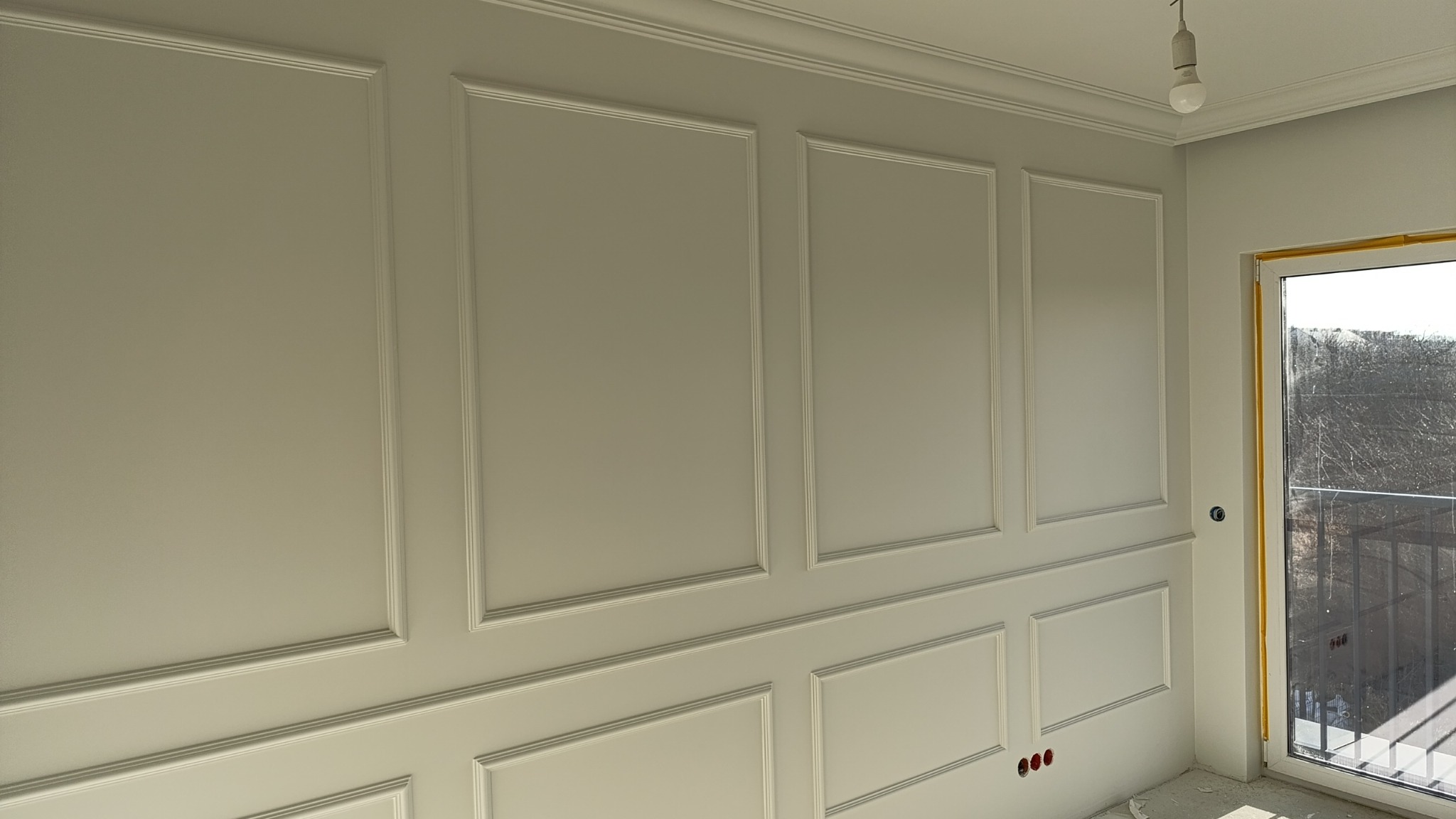 Ściana z dekoracyjnymi panelami w stylu klasycznym, pomalowana na jasny kolor. Widoczne listwy przypodłogowe i sufitowe, okno balkonowe z widokiem na zewnątrz.