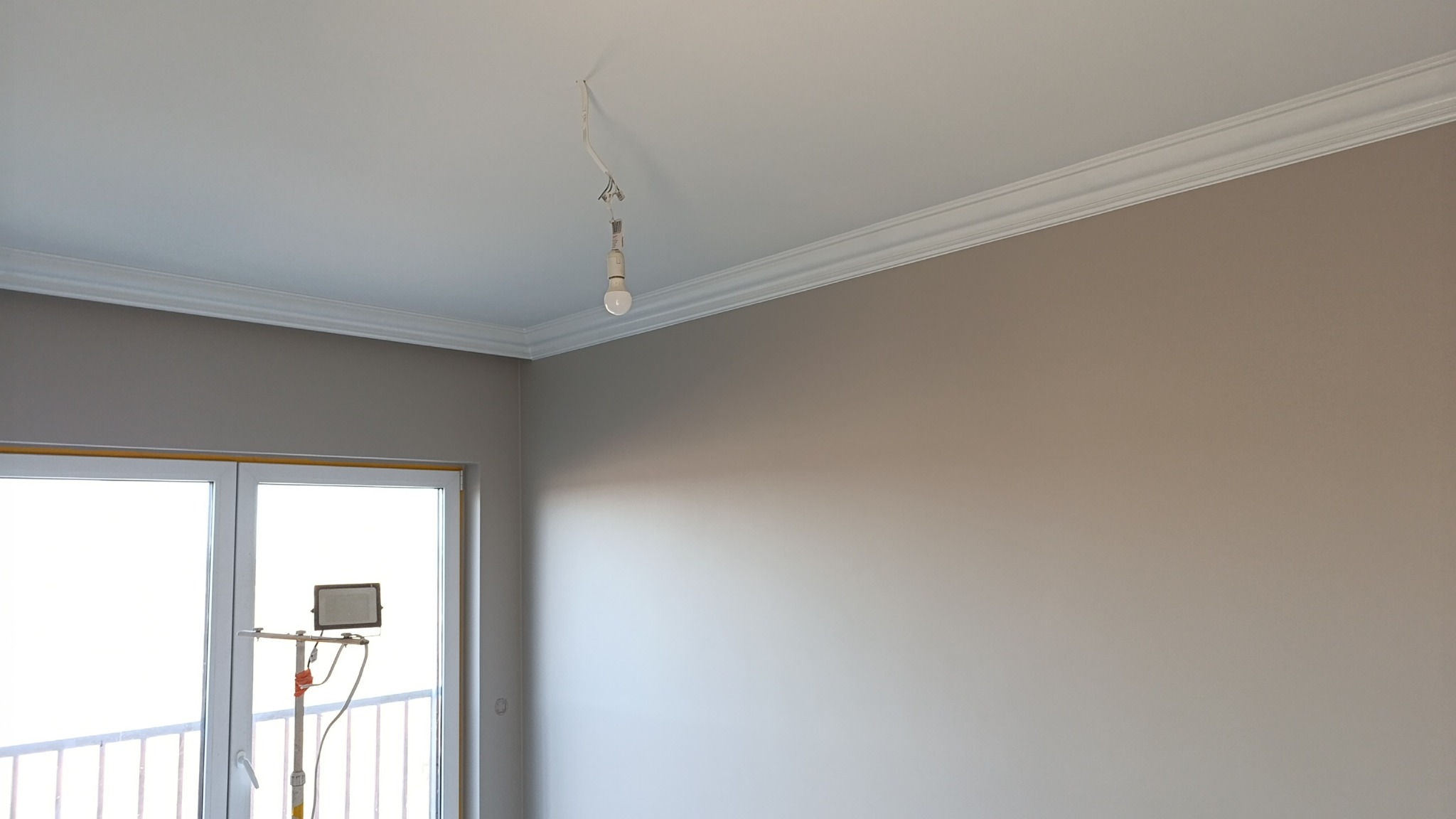 Pomieszczenie po malowaniu ścian na beżowo, z dekoracyjną listwą przysufitową i lampą wiszącą na kablu. Widoczne okno balkonowe i lampa oświetleniowa na statywie.