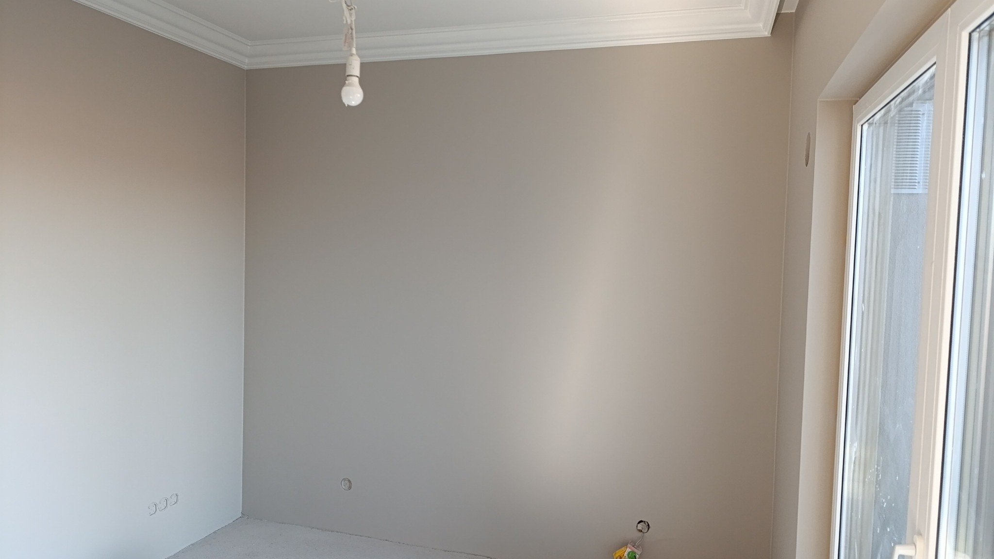 Świeżo pomalowane wnętrze pokoju z widocznymi gładkimi ścianami w odcieniach beżu i bieli, listwa przysufitowa i okno z roletami.