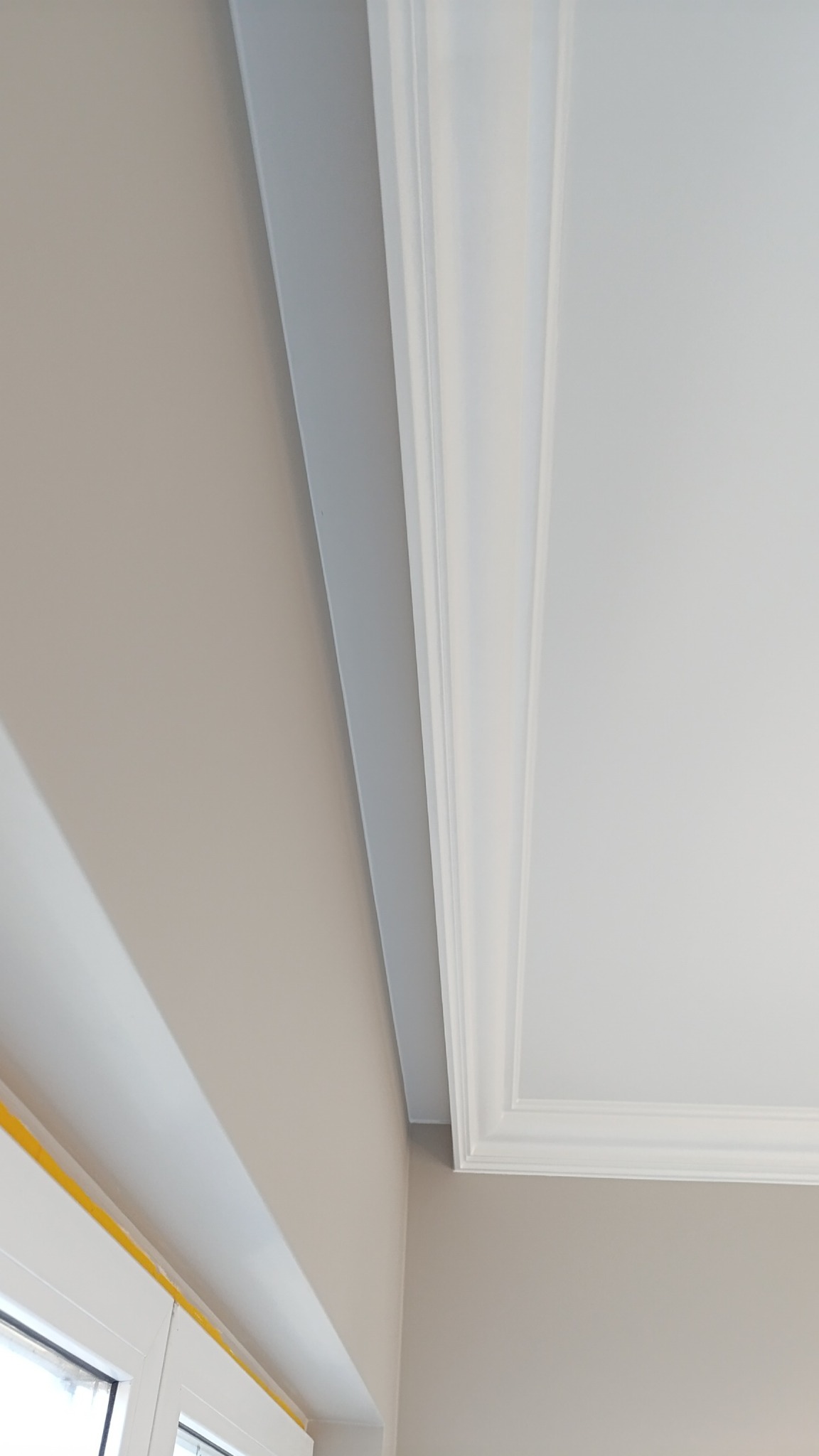 Detal wykończenia wnętrza: narożnik z listwą przysufitową i pomalowaną ścianą w neutralnym odcieniu beżu, zabezpieczone okno taśmą malarską.