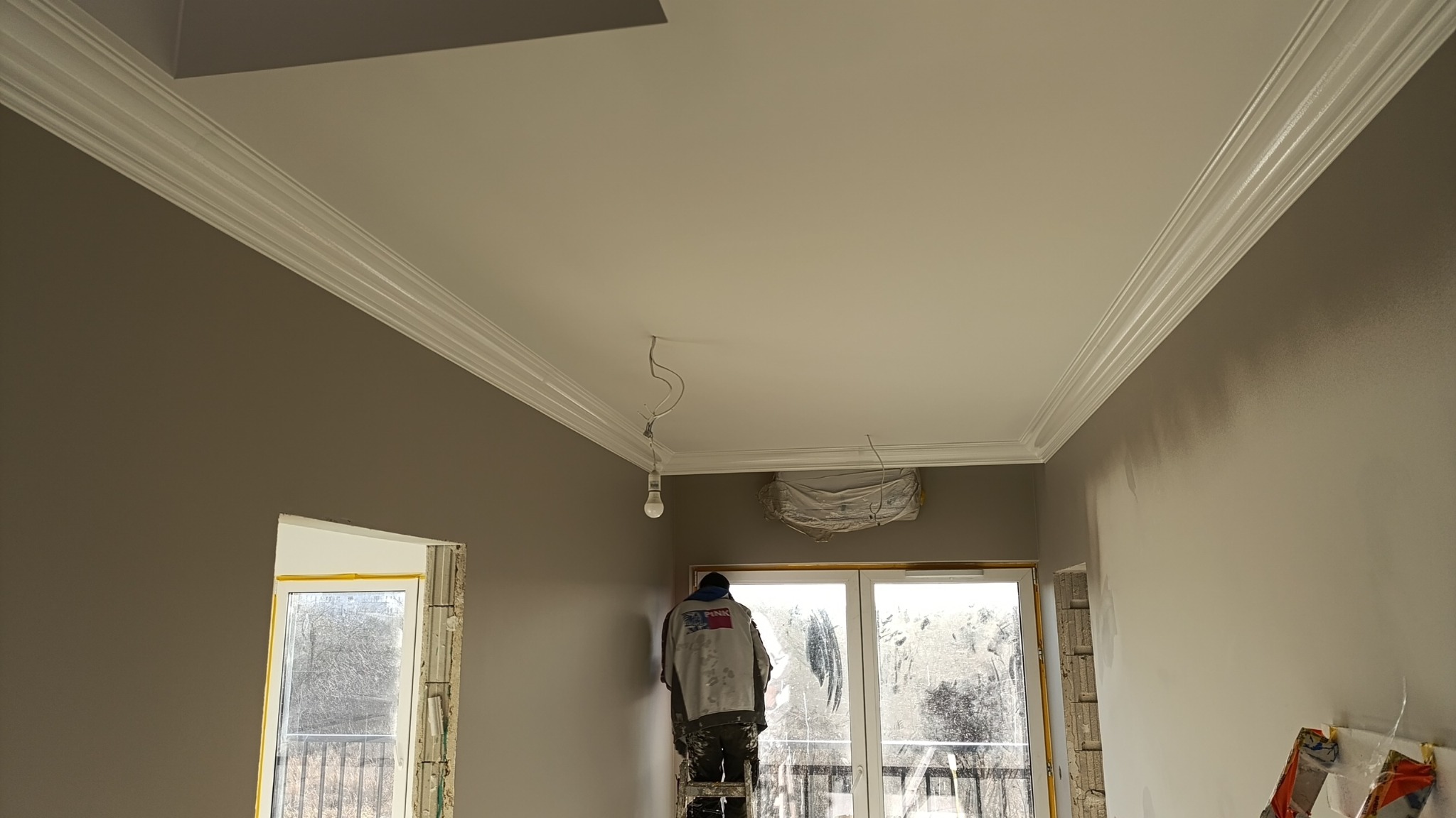Malarz na drabinie maluje ścianę w pokoju z ozdobną listwą sufitową. Widoczne okno balkonowe i fragmenty ścian w dwóch odcieniach szarości.