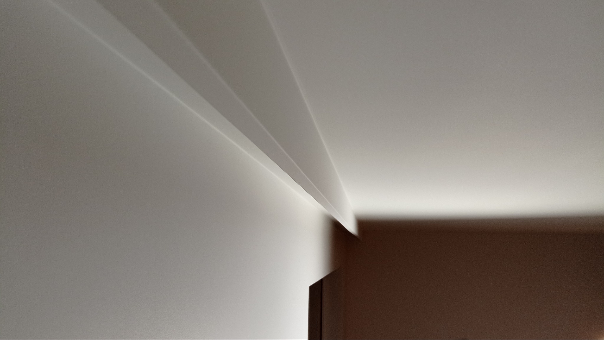 Detal wykończenia wnętrza: białe listwy przysufitowe zamontowane w rogu pokoju, oświetlone od dołu, tworzące minimalistyczny, nowoczesny efekt.