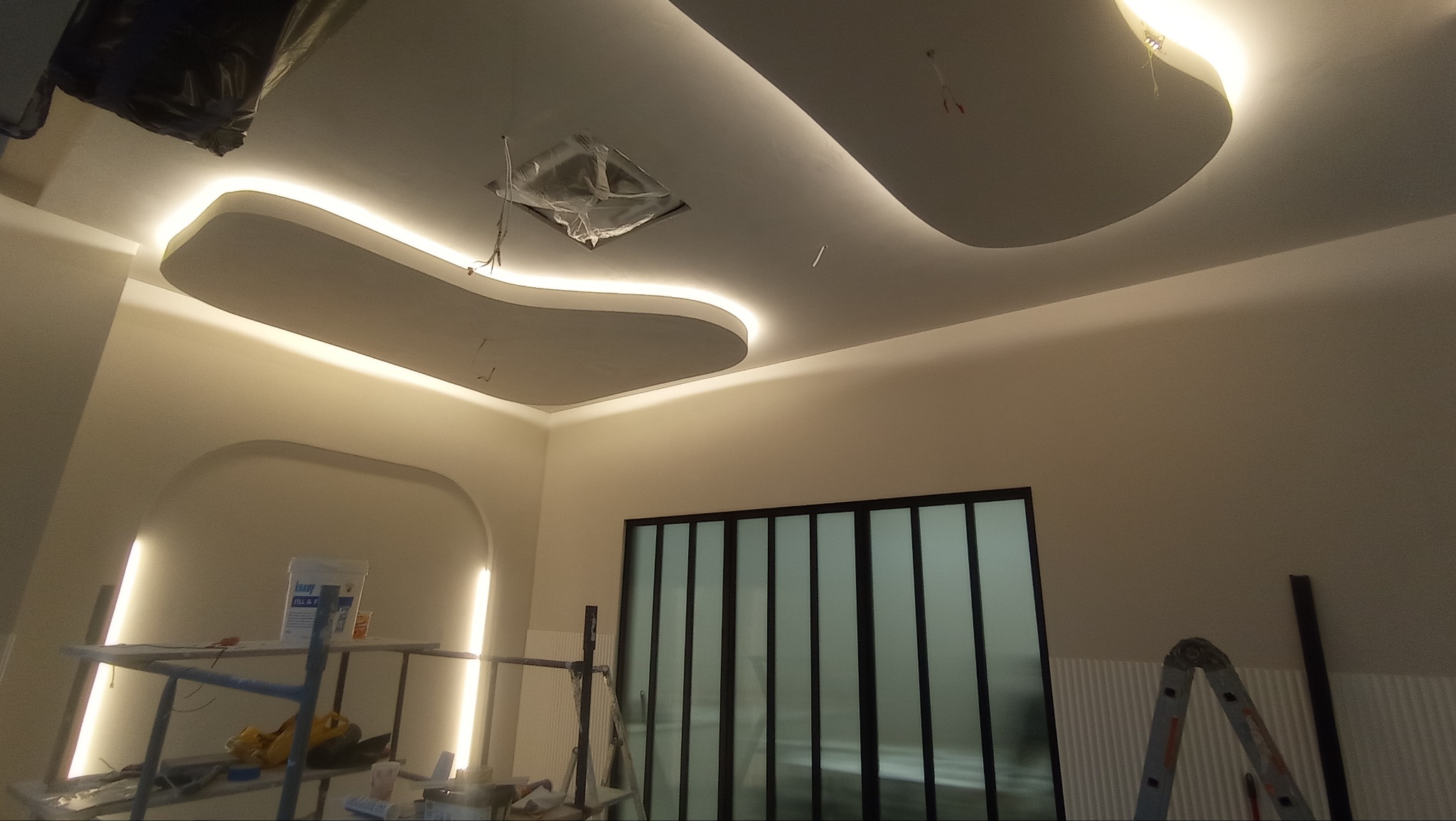 Nowoczesny sufit podwieszany z oświetleniem LED w kształcie fal, widok z dołu. W tle ściana i okno. Na pierwszym planie rusztowanie i drabina.