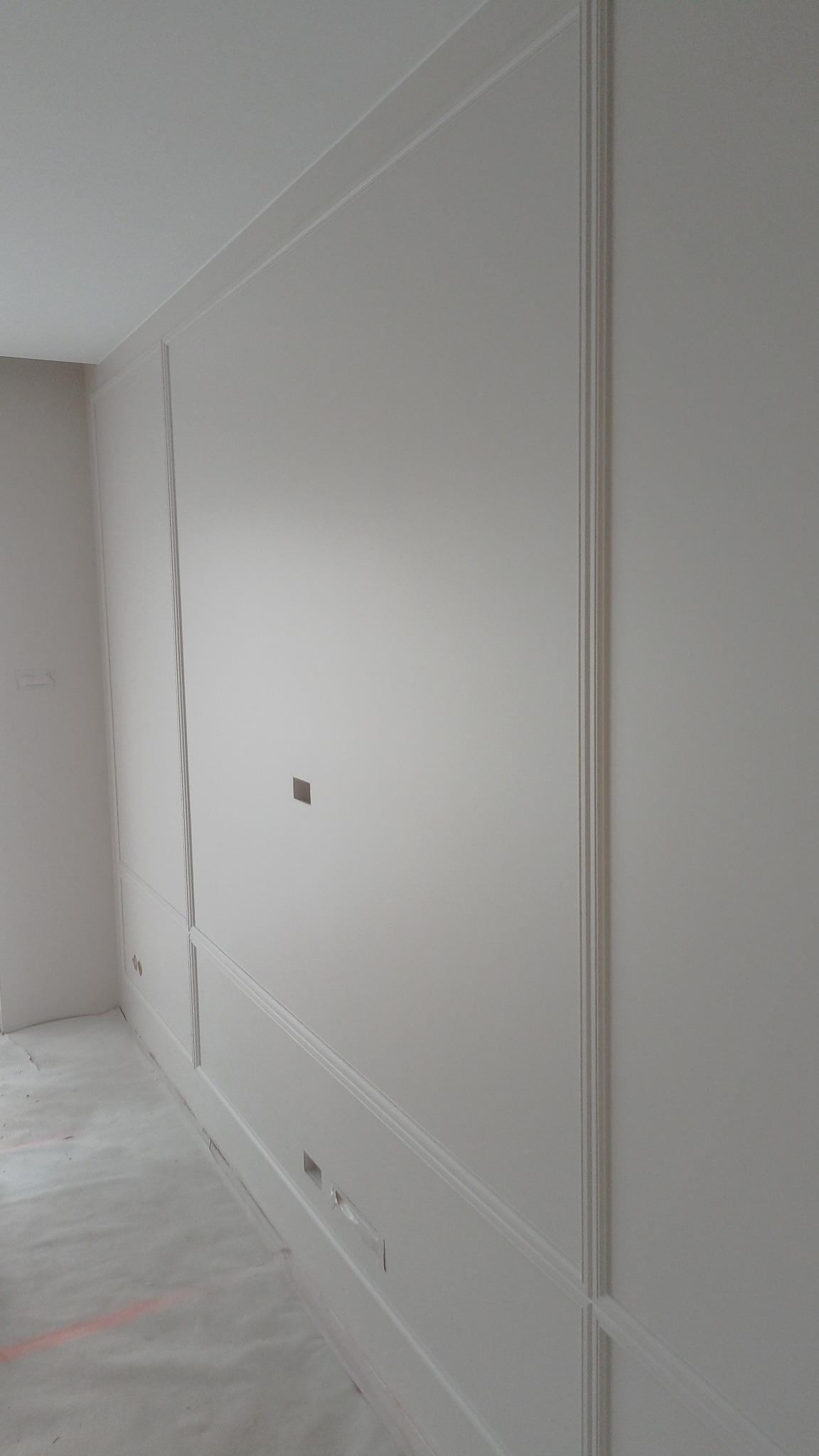 Wykończona ściana z dekoracyjnymi listwami w jasnym odcieniu, widoczne gniazdka elektryczne i otwór na przewody. Ściana świeżo po malowaniu, na podłodze folia ochronna.