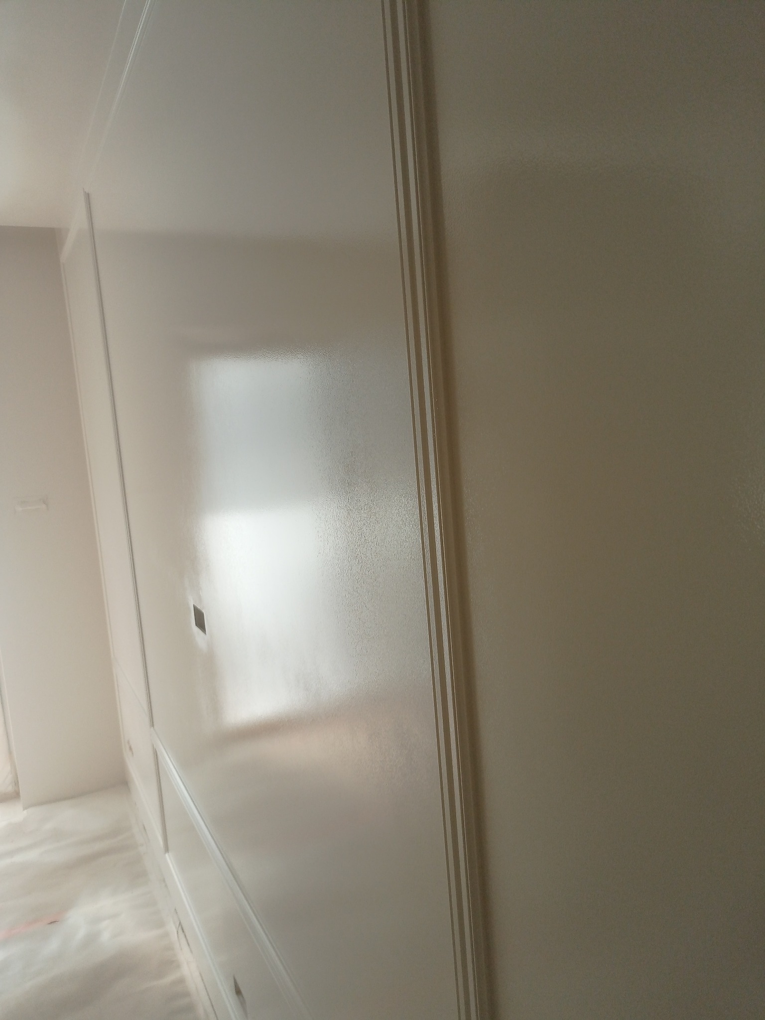 Biała, świeżo pomalowana ściana z dekoracyjnymi listwami, widoczna pod lekkim kątem. Na podłodze folia ochronna. Gładka powierzchnia, odbijające światło wykończenie.