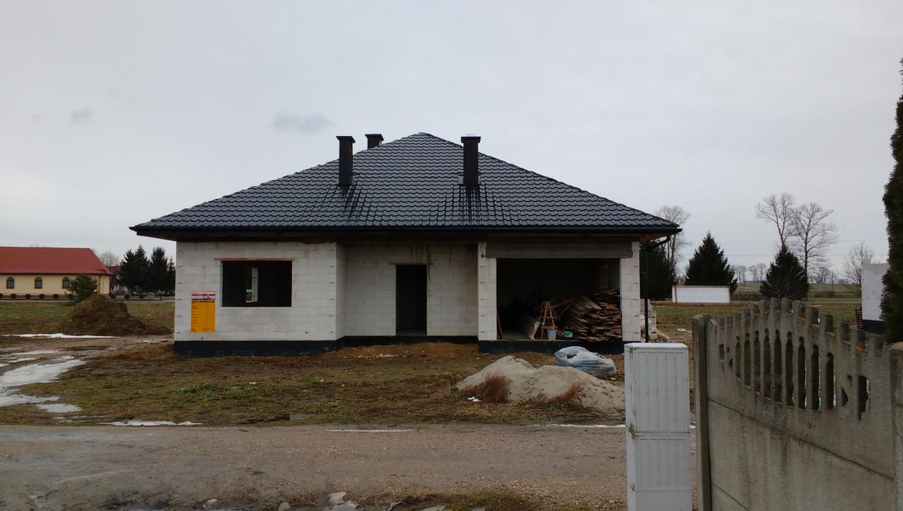 Budynek jednorodzinny w trakcie budowy, z widoczną konstrukcją z bloczków, czarnym dachem z dachówki, stertą drewna w garażu i ogłoszeniem na ścianie, na tle pochmurnego nieba.