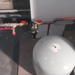 Instalacja hydrauliczna z widocznymi zaworami z czerwonymi pokrętłami, filtrem i zbiornikami wyrównawczymi, zamontowana w piwnicy lub kotłowni.