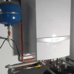 Nowoczesny, biały piec gazowy marki Vaillant zamontowany na ścianie, z widocznymi miedzianymi rurami, niebieskim zbiornikiem wyrównawczym i elementami instalacji podłączeniowej.