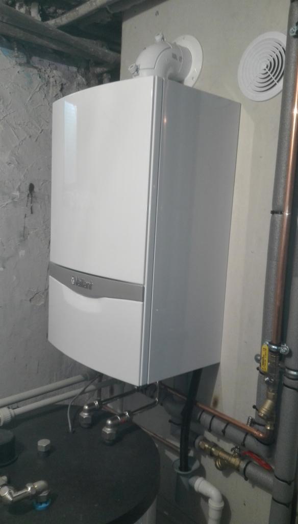 Biały, kondensacyjny piec gazowy marki Vaillant zamontowany na ścianie z widocznymi rurami miedzianymi i elementami instalacji, w tle surowa ściana z odsłoniętym betonem.