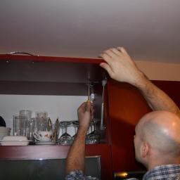 BenySerwis - Mężczyzna dokręca śrubokrętem amortyzator podnośnika w szafce kuchennej, widoczne naczynia i szkło wewnątrz szafki.