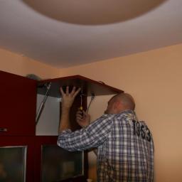 BenySerwis - Mężczyzna w koszuli w kratę montuje szafkę kuchenną w Bielanach Wrocławskich, używając śrubokręta; widoczne siłowniki gazowe podtrzymujące drzwiczki.