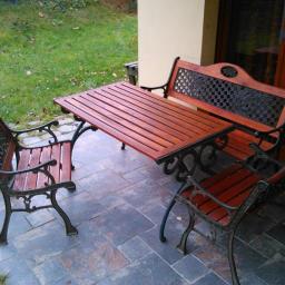 BenySerwis - Zestaw mebli ogrodowych: stół z drewnianym blatem i dwie ławki z metalowymi zdobieniami, ustawione na kamiennej posadzce tarasu.