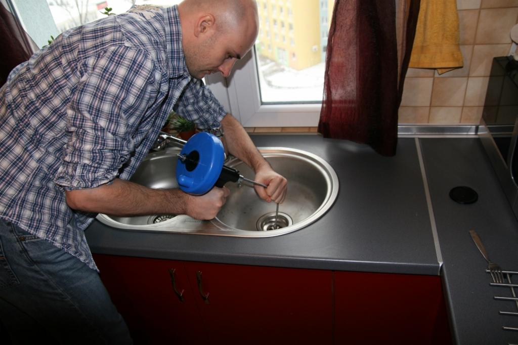 Mężczyzna w koszuli w kratę używa niebieskiego przepychacza sprężynowego do odblokowania zatkanego odpływu w zlewie kuchennym.