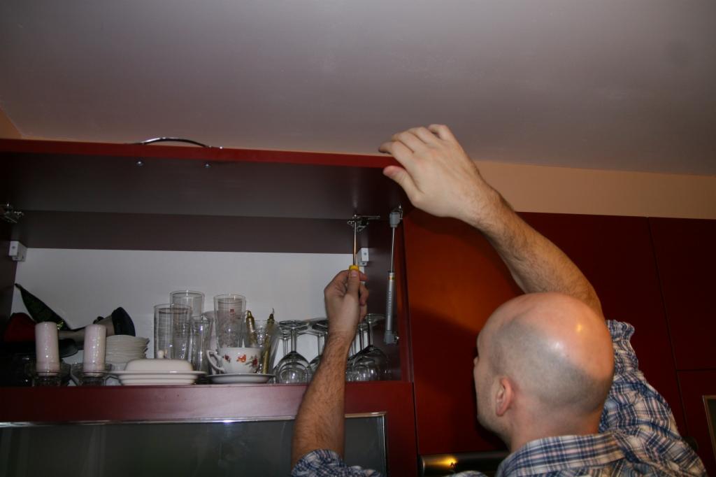 Mężczyzna dokręca śrubokrętem amortyzator podnośnika w szafce kuchennej, widoczne naczynia i szkło wewnątrz szafki.