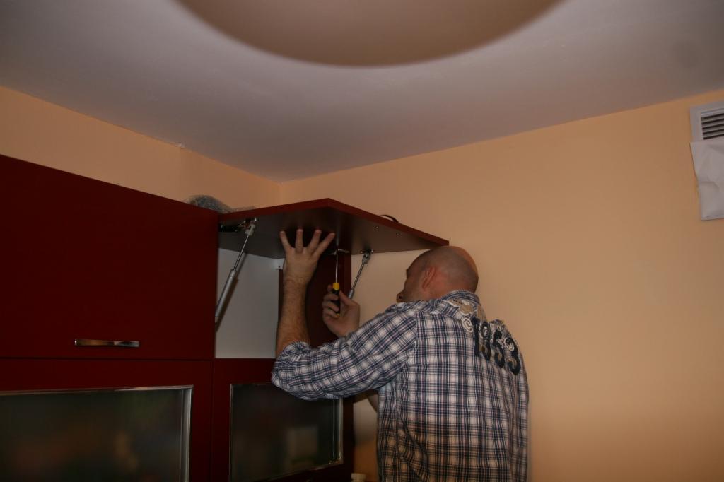 Mężczyzna w koszuli w kratę montuje szafkę kuchenną w Bielanach Wrocławskich, używając śrubokręta; widoczne siłowniki gazowe podtrzymujące drzwiczki.