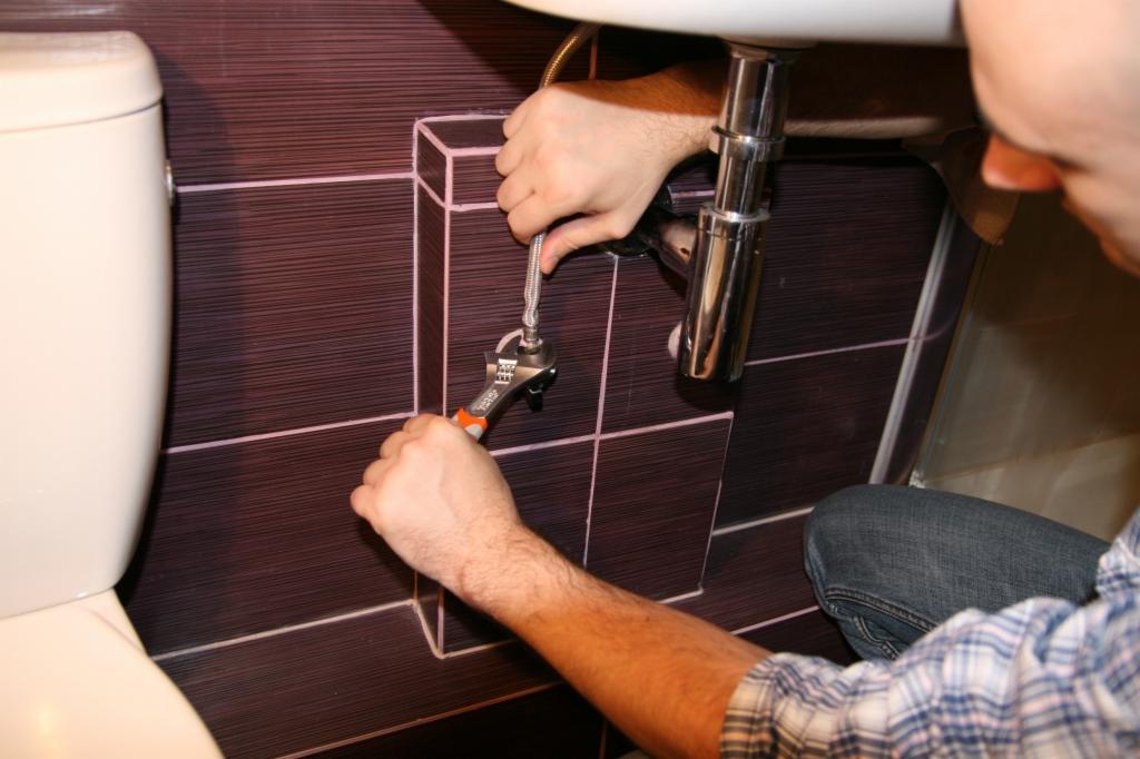 Instalator dokręca elastyczny wężyk doprowadzający wodę do umywalki za pomocą klucza nastawnego; widoczne ciemne płytki z jasnymi fugami i fragment toalety.