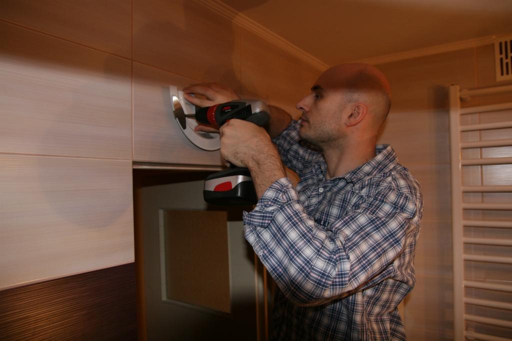 Mężczyzna w koszuli w kratę wierci w płytkach ściennych w łazience, montując biały uchwyt, używając wiertarki akumulatorowej.