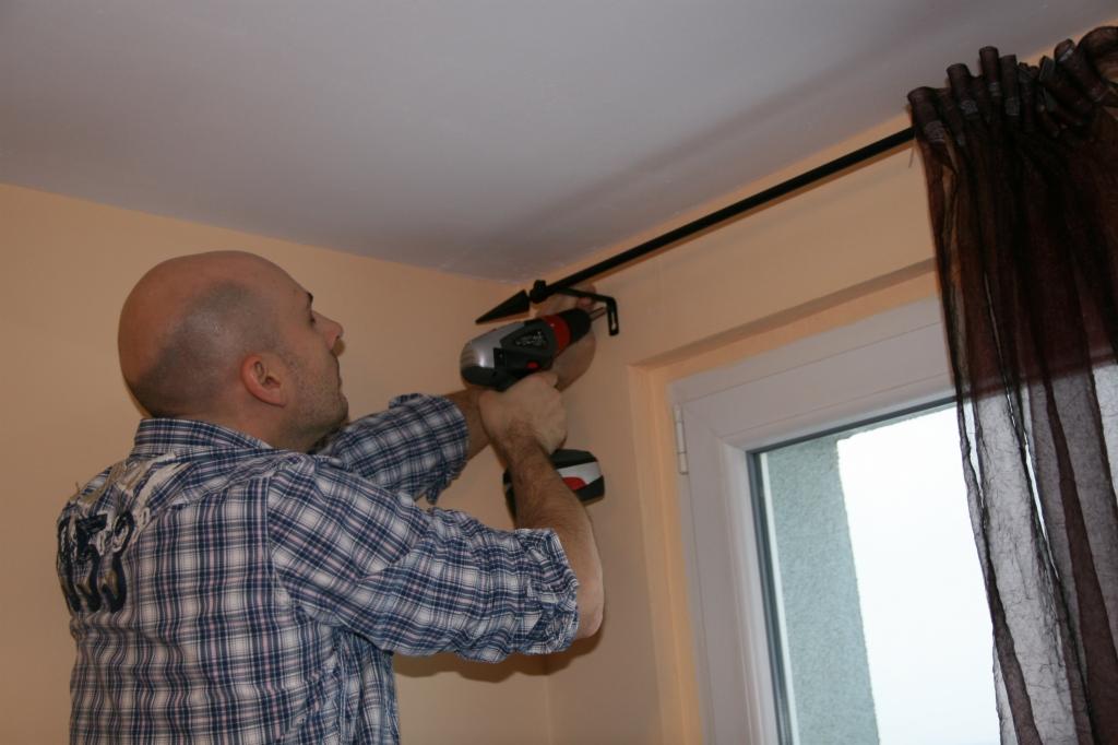 Mężczyzna w koszuli w kratę montuje karnisz nad oknem za pomocą wkrętarki akumulatorowej.