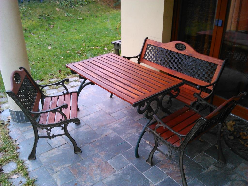 Zestaw mebli ogrodowych: stół z drewnianym blatem i dwie ławki z metalowymi zdobieniami, ustawione na kamiennej posadzce tarasu.