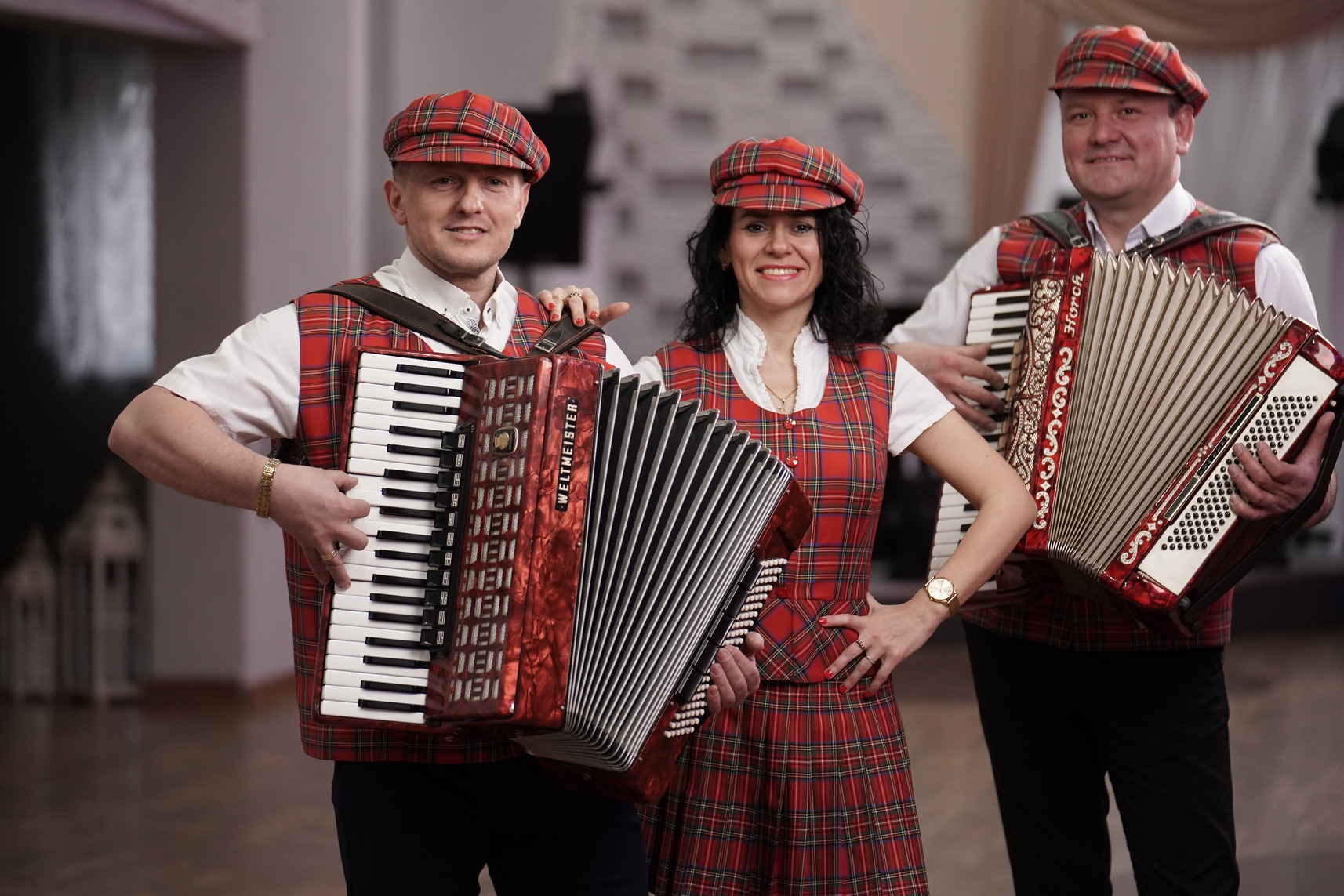 Trzyosobowy zespół w czerwono-czarną kratę prezentuje się z akordeonami Weltmeister i Horch, uśmiechnięci i gotowi do występu.