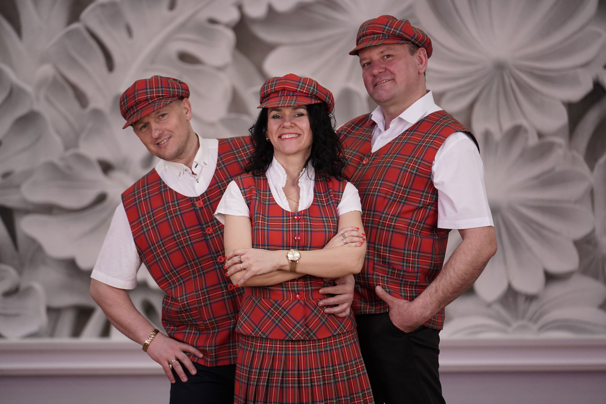 Trzyosobowy zespół w dopasowanych strojach w szkocką kratę: kapelusze, kamizelki i spódnica. Członkowie zespołu uśmiechają się, pozując na tle ściany z motywem kwiatowym.