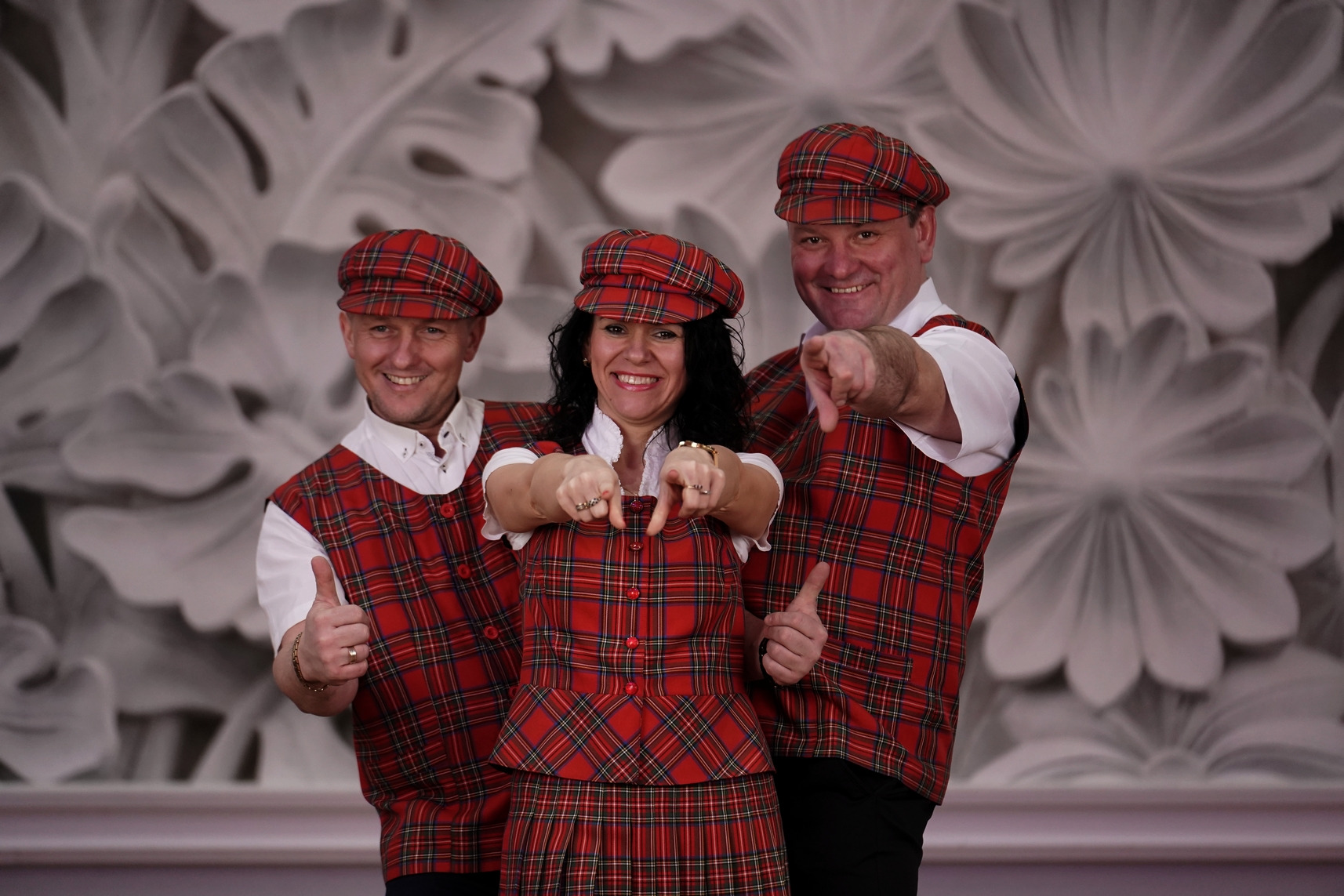 Trzy osoby w czerwono-czarnej szkockiej kracie, uśmiechnięte i wskazujące palcami na widza, z dekoracyjnym tłem w kwiaty.