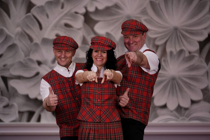 Trzyosobowy zespół w czerwono-czarną kratę, w kamizelkach i kaszkietach, uśmiechnięci, wskazują palcami w stronę widza, na tle ściany z motywem kwiatowym.