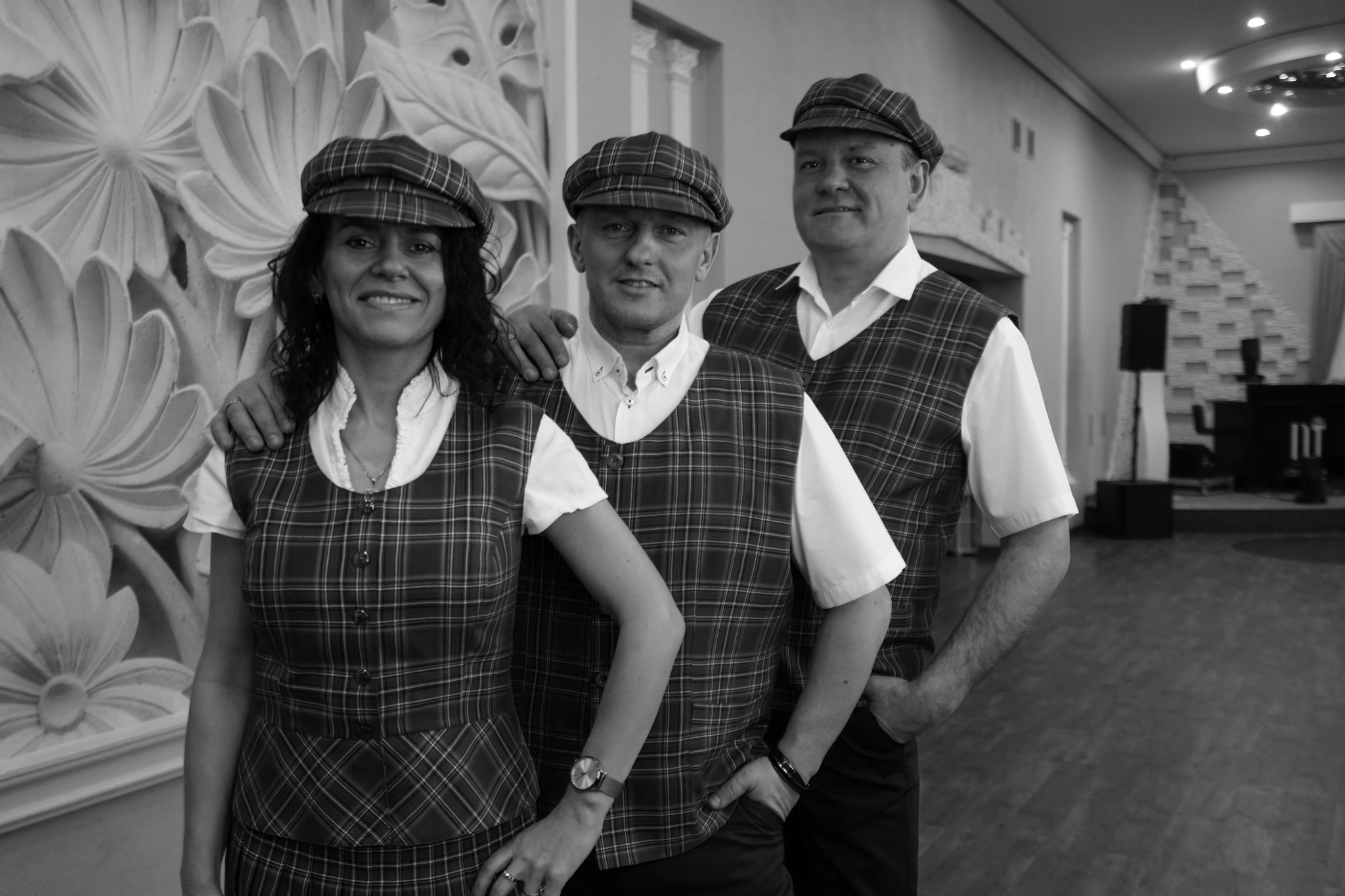 Zespół muzyczny w kratowanych kamizelkach i kaszkietach pozuje na tle ściany z kwiatowym ornamentem w sali bankietowej, widoczny fragment sceny z instrumentami.