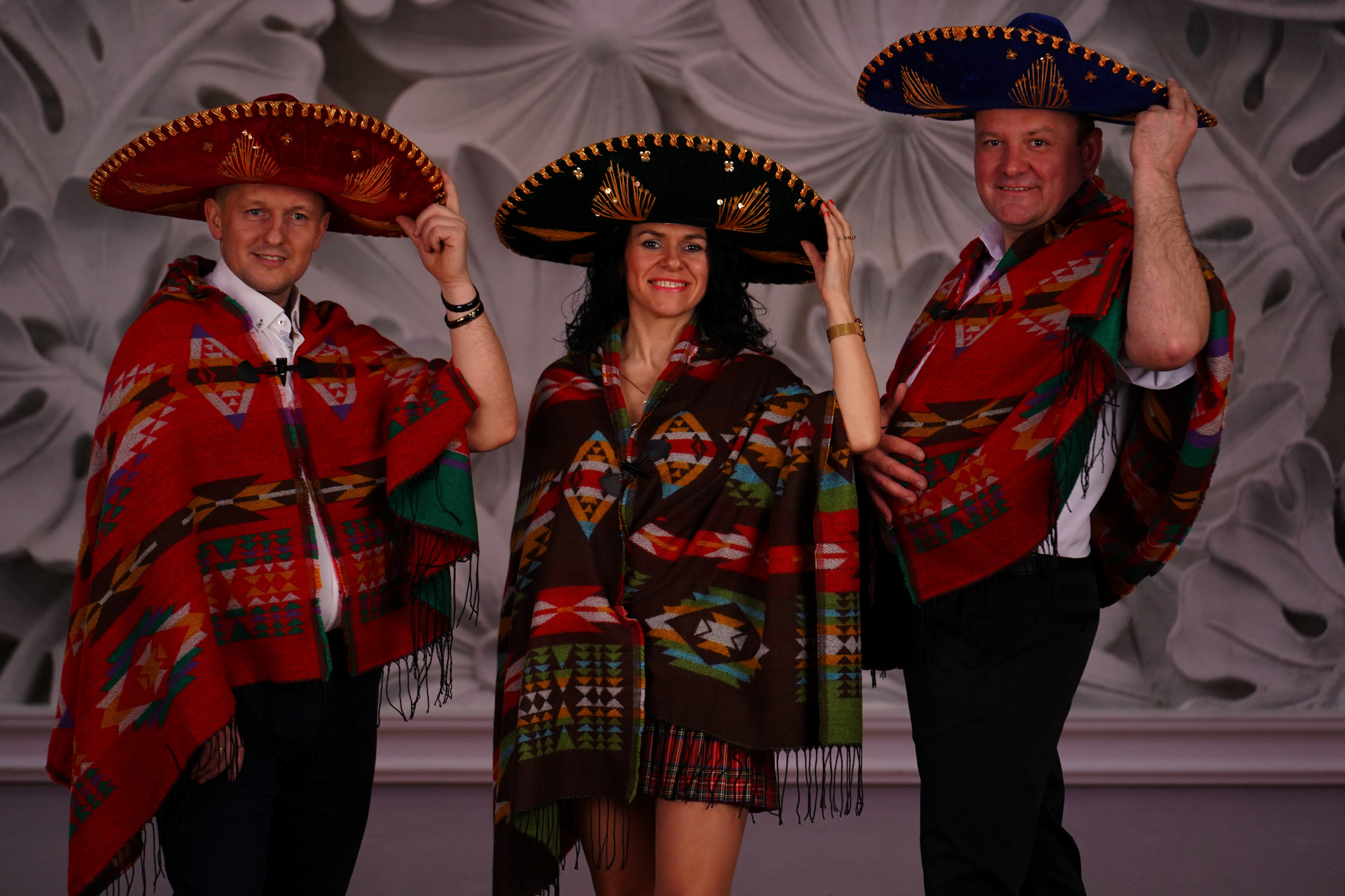 Trzy osoby w strojach meksykańskich: mężczyźni w czerwonych ponczach i sombrerach, kobieta w brązowym ponczo i zielonym sombrero, wszyscy uśmiechnięci na tle dekoracyjnej ściany.