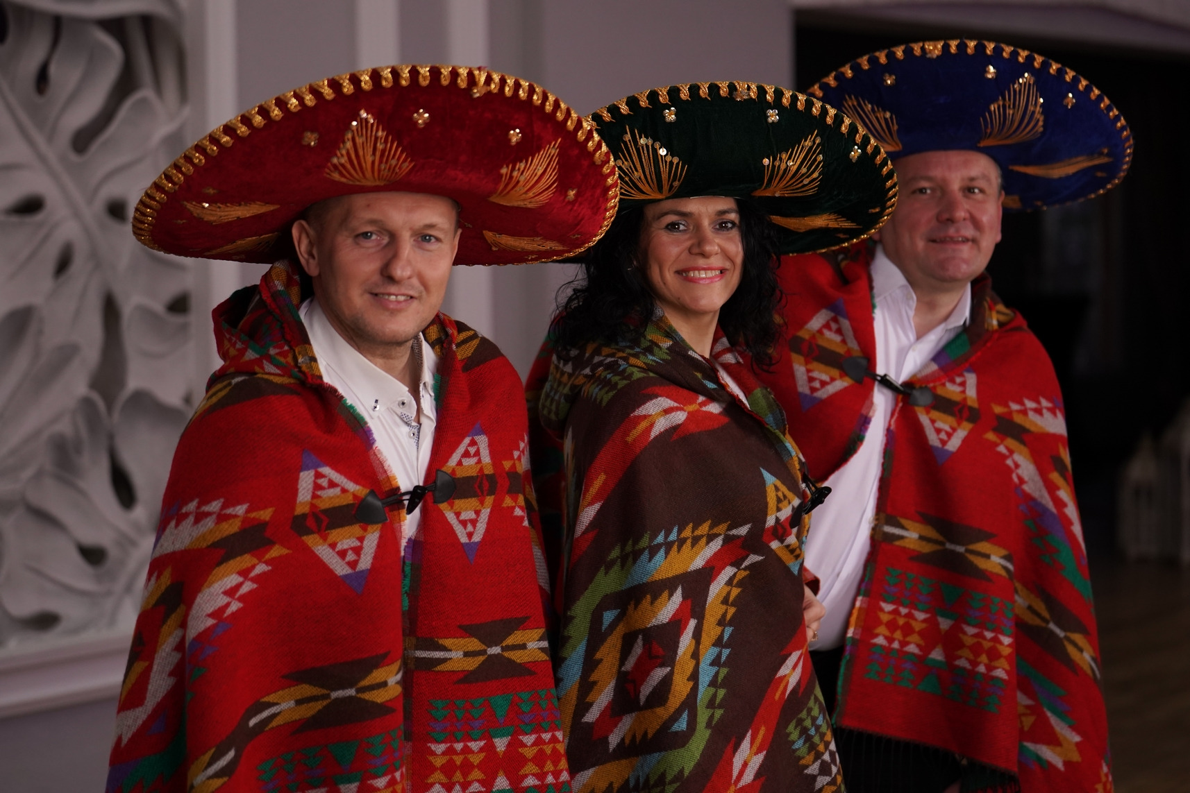 Trzy osoby w kolorowych sombrerach i ponczach z etnicznymi wzorami, pozujące do zdjęcia. Mężczyzna po lewej uśmiecha się, a jego ponczo jest czerwone. Kobieta pośrodku uśmiecha się, a jej ponczo...