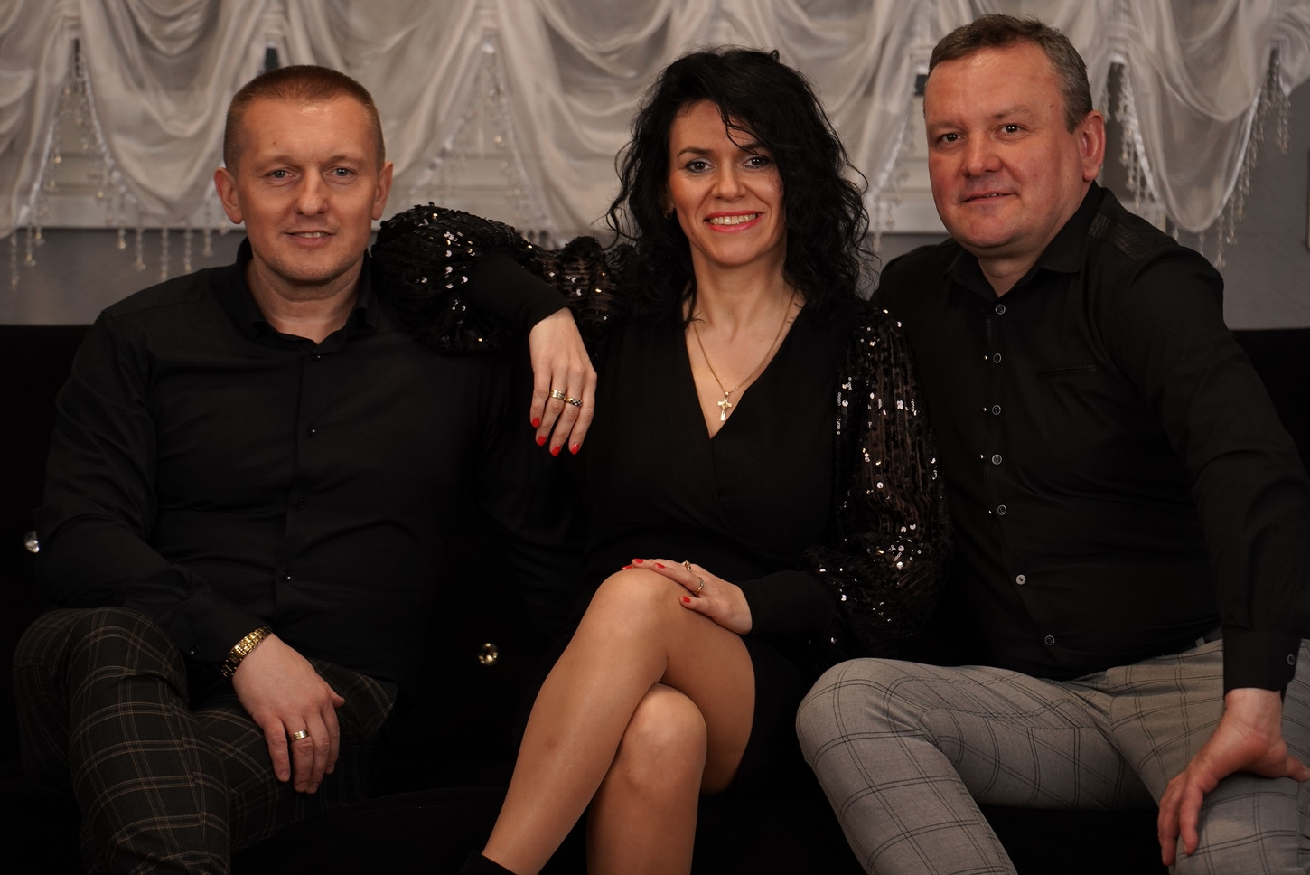 Trzyosobowy zespół w czarnych strojach pozuje na czarnej kanapie przed błyszczącą, białą zasłoną.