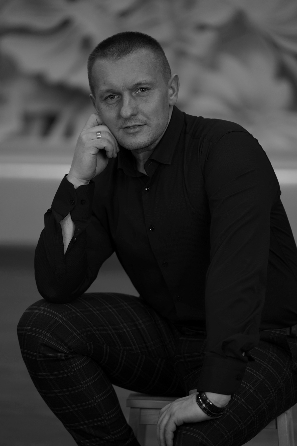 Portret mężczyzny w czarnej koszuli i spodniach w kratę, siedzącego na stołku, z ręką opartą o policzek, w czarno-białej tonacji.
