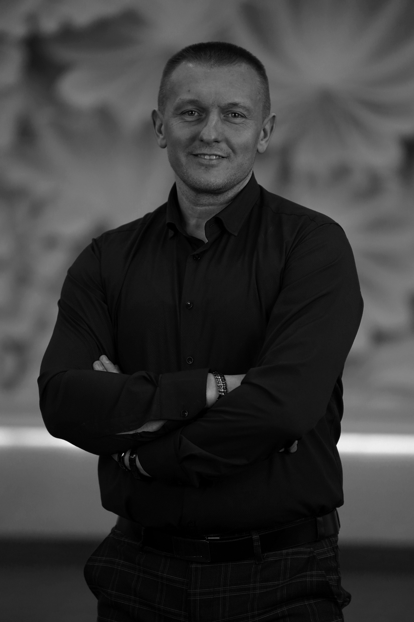 Portret mężczyzny w czarnej koszuli z długim rękawem, z założonymi rękami i bransoletką na nadgarstku, na rozmytym tle.