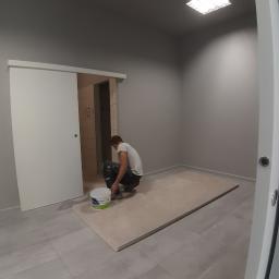 M.S. Vel-BUD Firma Rodzinna - Mężczyzna w szarym pomieszczeniu, klęczący obok wiadra z farbą, przygotowuje płyty kartonowo-gipsowe do montażu, widoczne drzwi przesuwne i szare ściany.